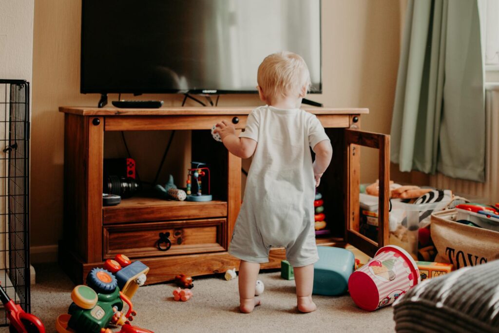 A toddler playing in a messy living room filled with colorful toys and a TV stand.
