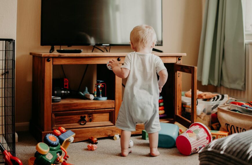A toddler playing in a messy living room filled with colorful toys and a TV stand.