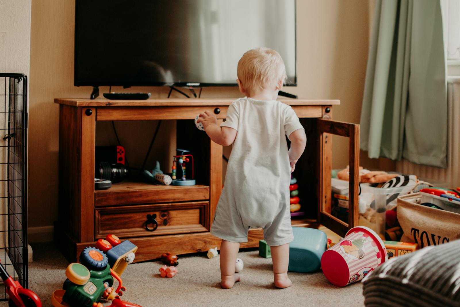 A toddler playing in a messy living room filled with colorful toys and a TV stand.