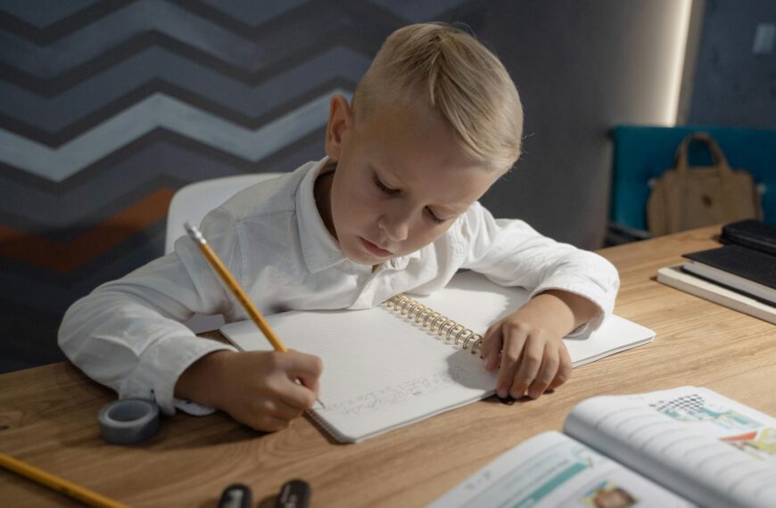 A young boy diligently writing homework in a notebook at a wooden desk indoors.