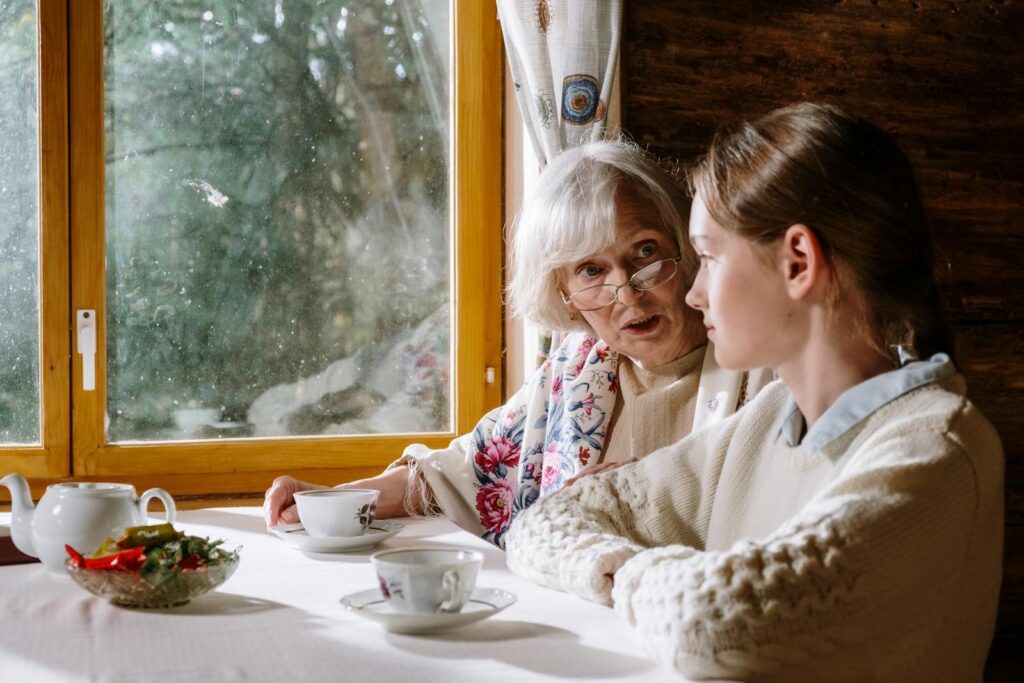 Warm indoor scene of a grandmother and granddaughter sharing tea and conversation by a window.