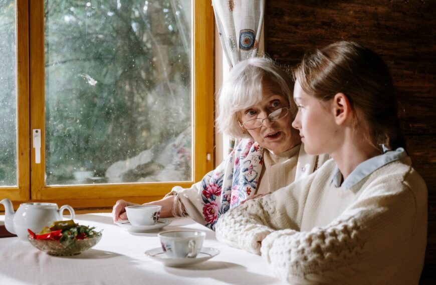 Warm indoor scene of a grandmother and granddaughter sharing tea and conversation by a window.