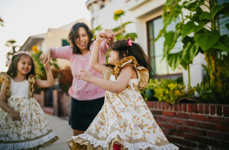 A mother dances with her twin daughters in a lively outdoor setting, enjoying a sunny day.