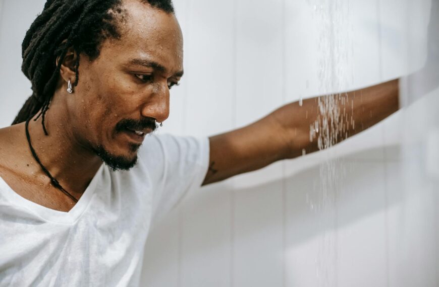 Side view of crop young worried African American man with dreadlocks in white t shirt leaning hand on tiled wall while standing under water stream in bathroom