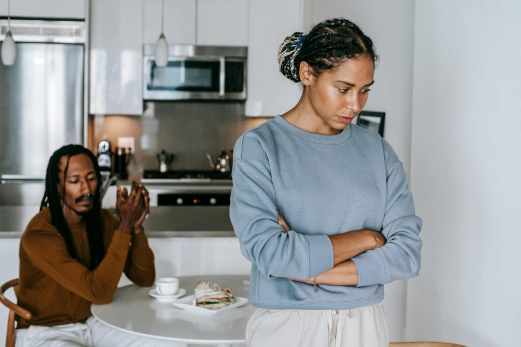 Young discontented African American female with folded arms against male partner at kitchen table during quarrel