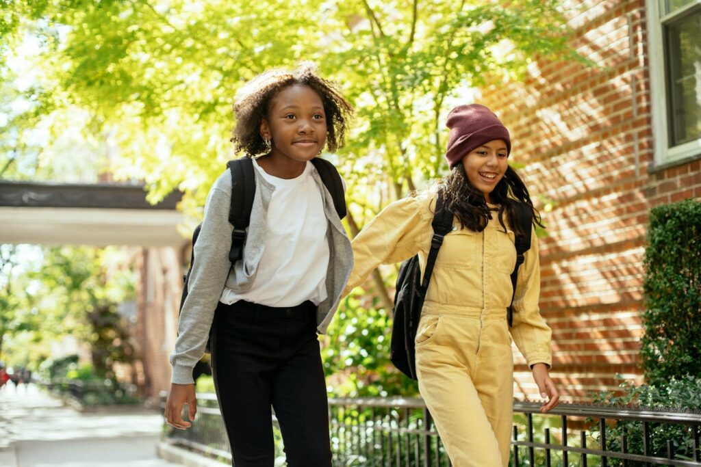 Two cheerful girls walking hand-in-hand outdoors under sunny skies, enjoying a school day.