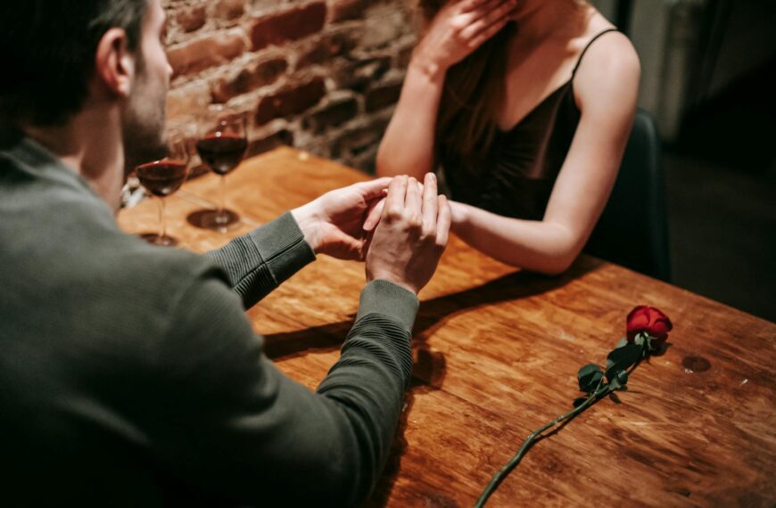 Crop unrecognizable couple in elegant clothes sitting in cafeteria at table with wine in glasses and rose near brick wall while having romantic dinner and holding hand while making proposal