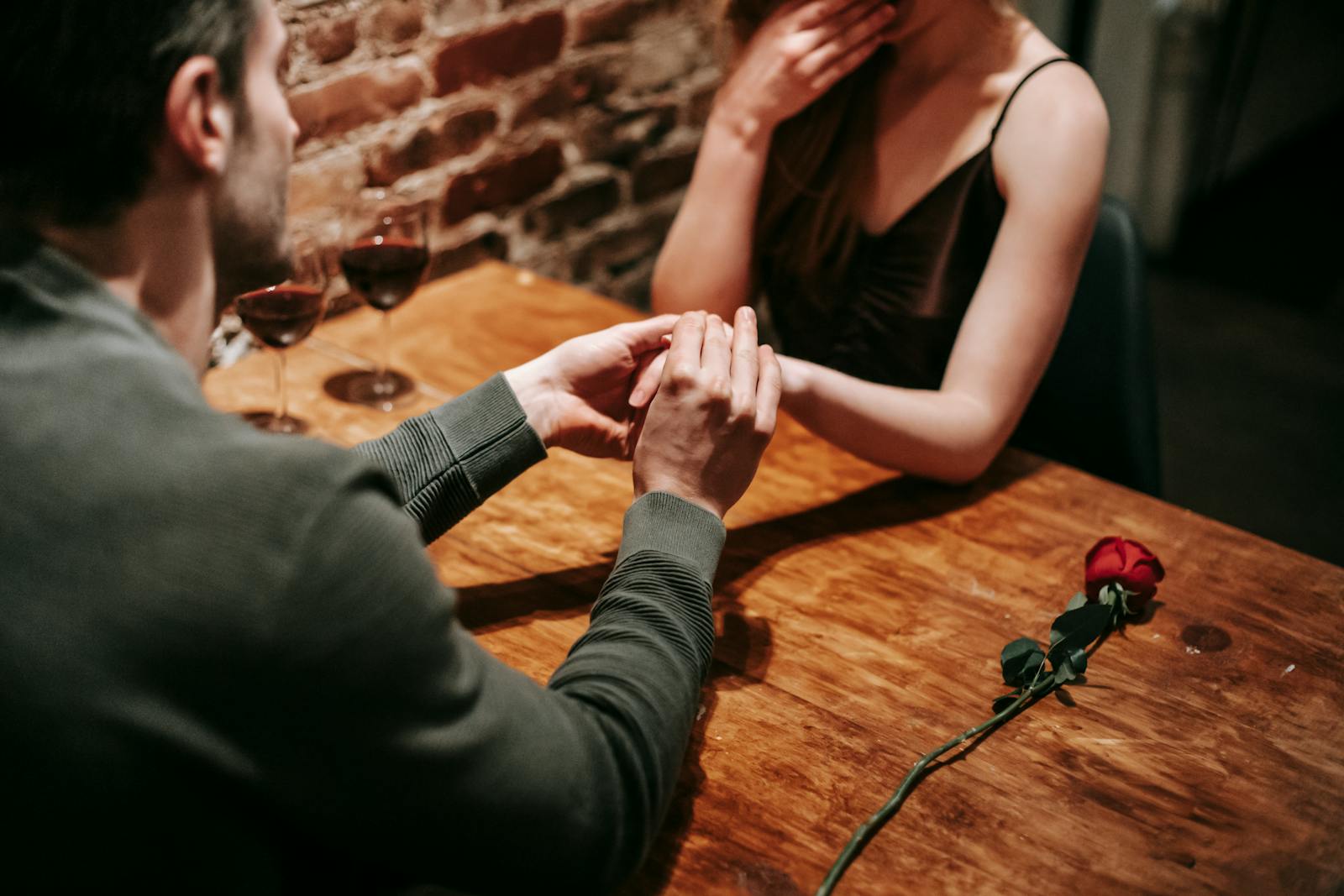 Crop unrecognizable couple in elegant clothes sitting in cafeteria at table with wine in glasses and rose near brick wall while having romantic dinner and holding hand while making proposal