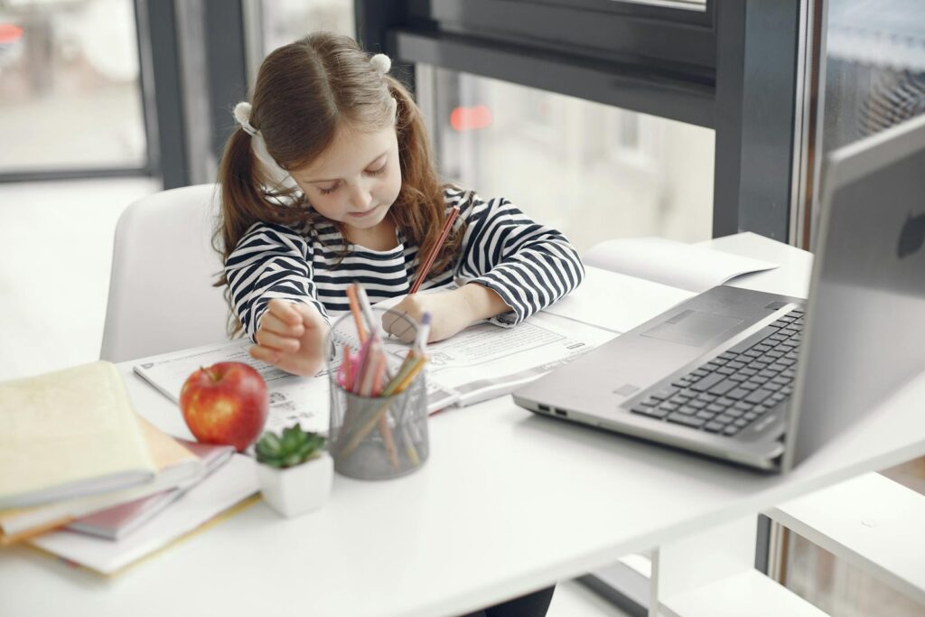 A young girl studies at home using a laptop, engaged with her homework and school supplies.
