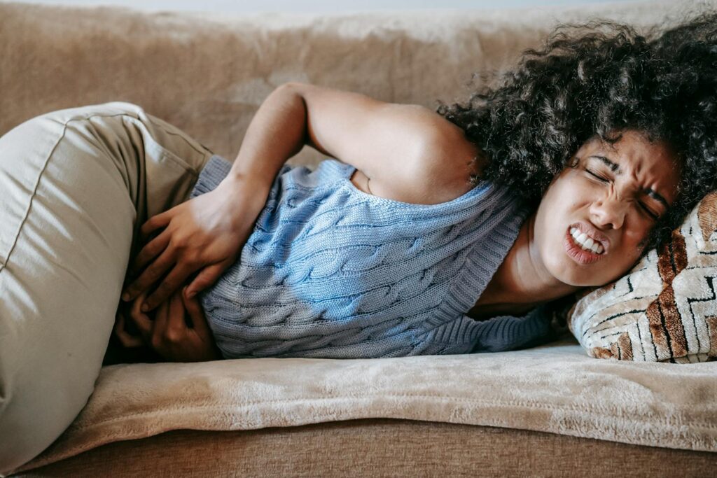 A woman lying on a couch, visibly in pain from stomach cramps, reflecting symptoms of menstruation or indigestion.
