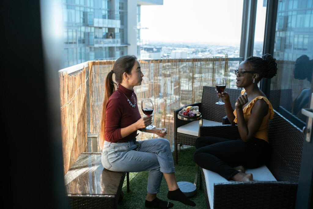 Two women enjoying wine and conversation on a city balcony with a scenic view.