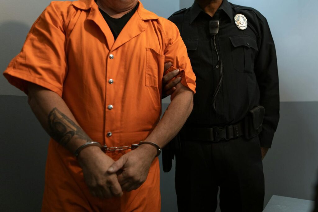 A handcuffed inmate in orange prison uniform being escorted by a police officer.