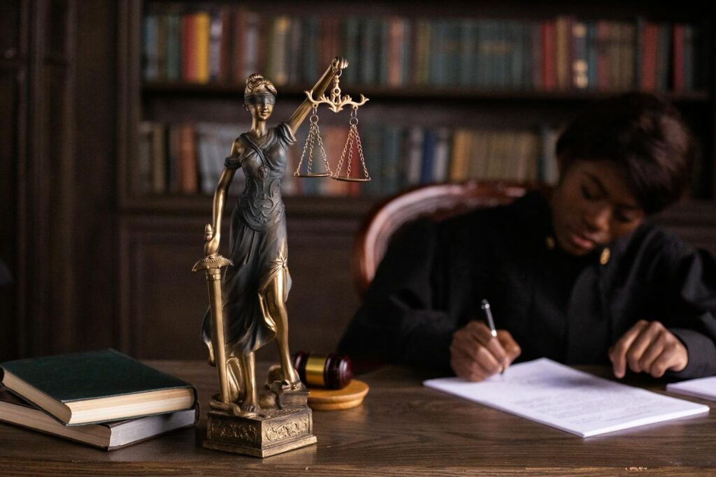 A focused judge writing on documents beside a Lady Justice statue in an office.