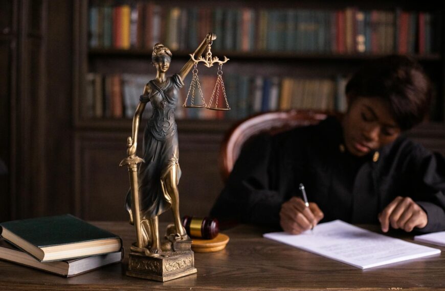 A focused judge writing on documents beside a Lady Justice statue in an office.