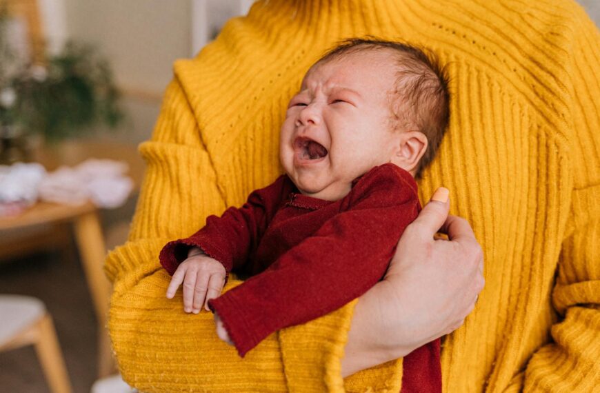 Emotional portrait of a crying newborn being comforted in caregiver's arms indoors.