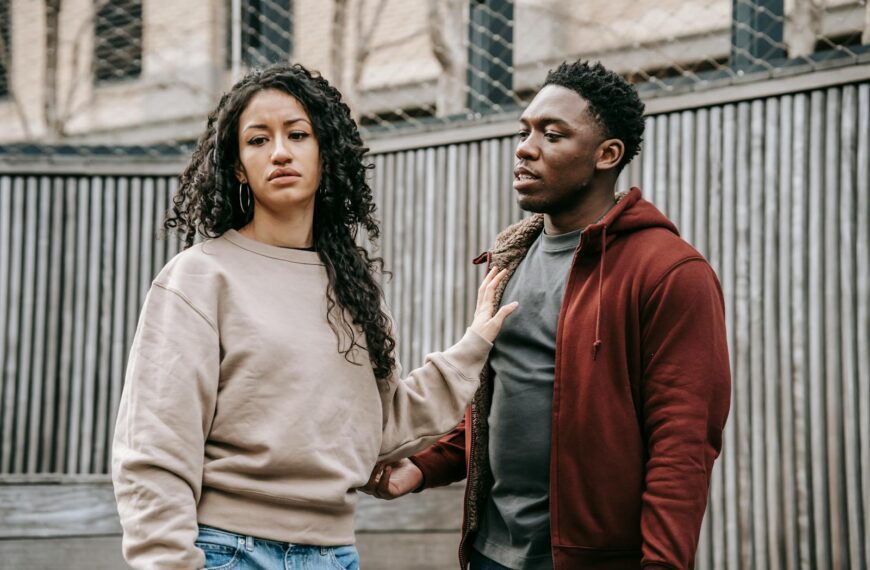 Sad ethnic girlfriend with curly hair rejecting annoyed African American boyfriend while arguing on street near wooden fence during breakup