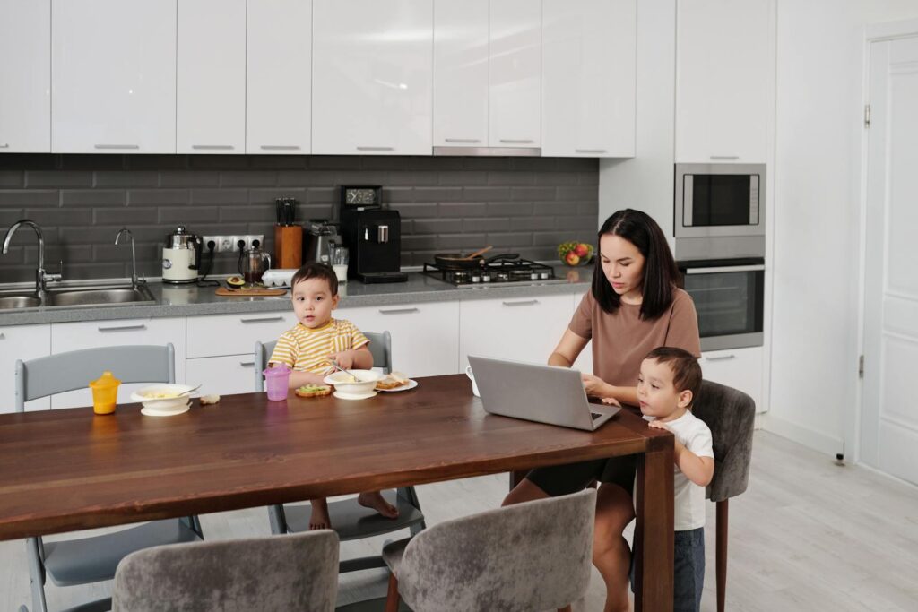 A mother works on her laptop while her children eat breakfast at the kitchen table.
