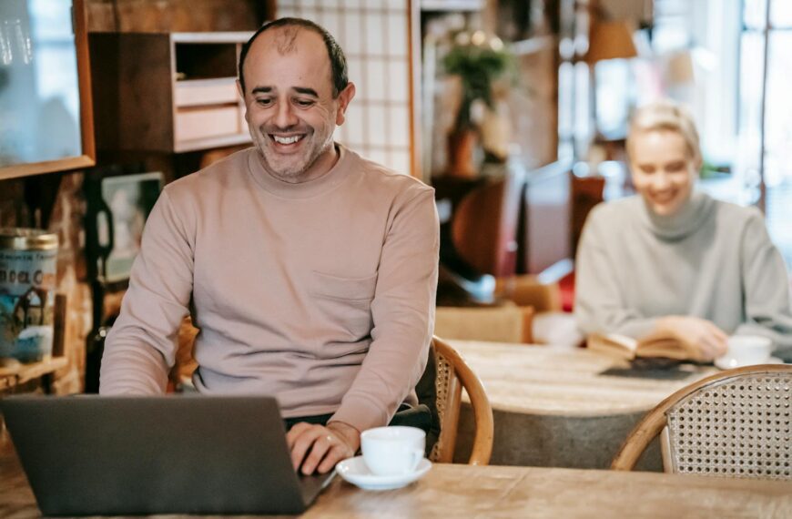 Cheerful middle aged ethnic man smiling while working remotely on laptop near positive wife reading book and drinking coffee