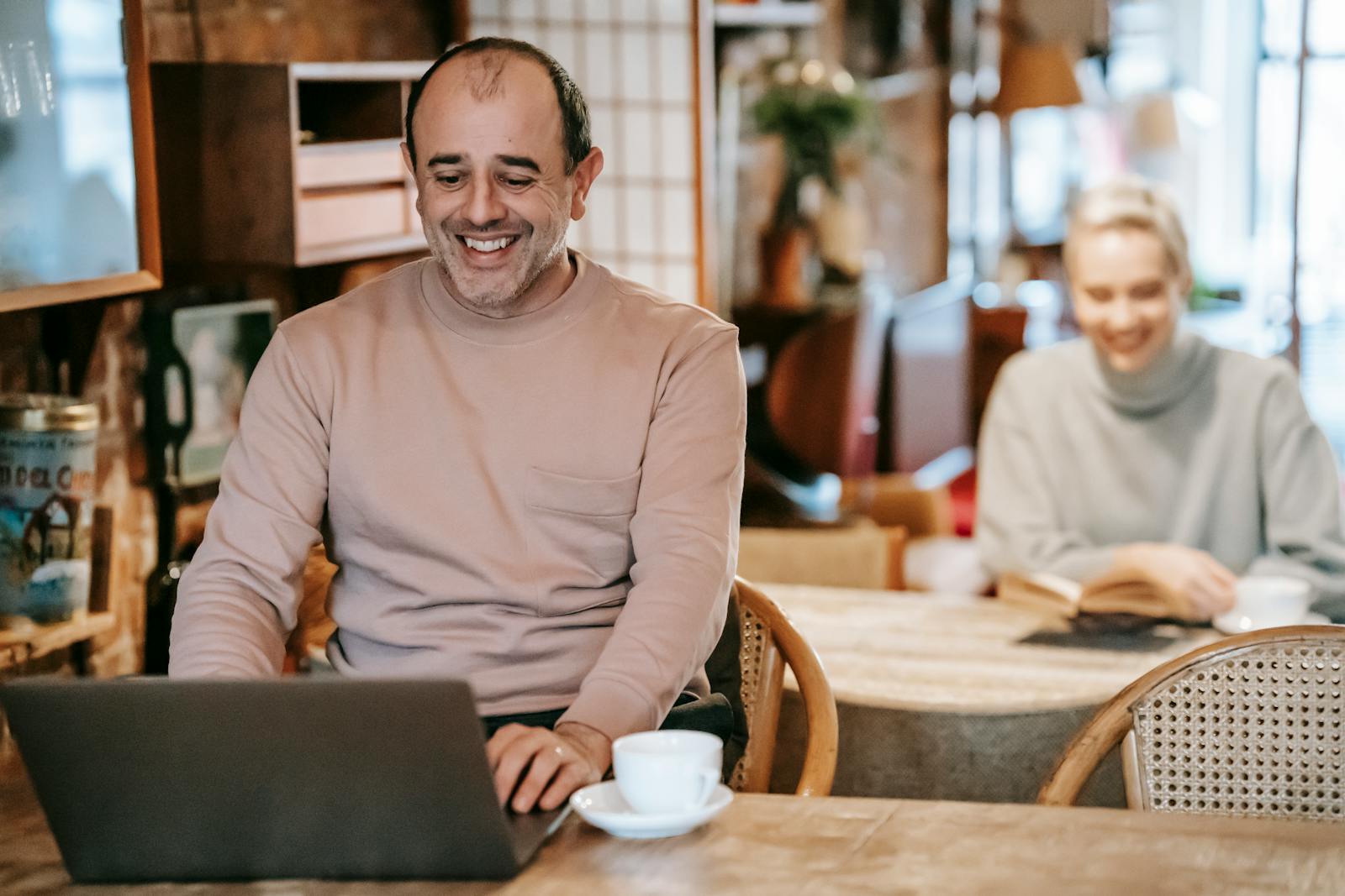 Cheerful middle aged ethnic man smiling while working remotely on laptop near positive wife reading book and drinking coffee