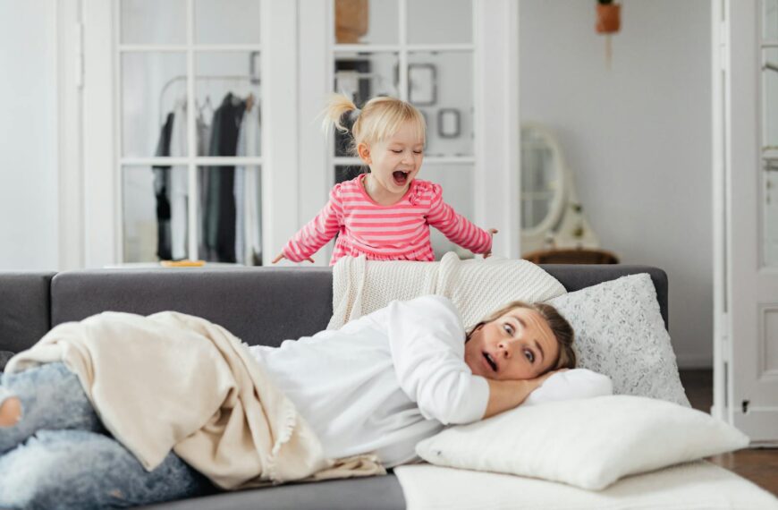 A young girl playfully shouting behind a resting mother on a couch in a cozy room.