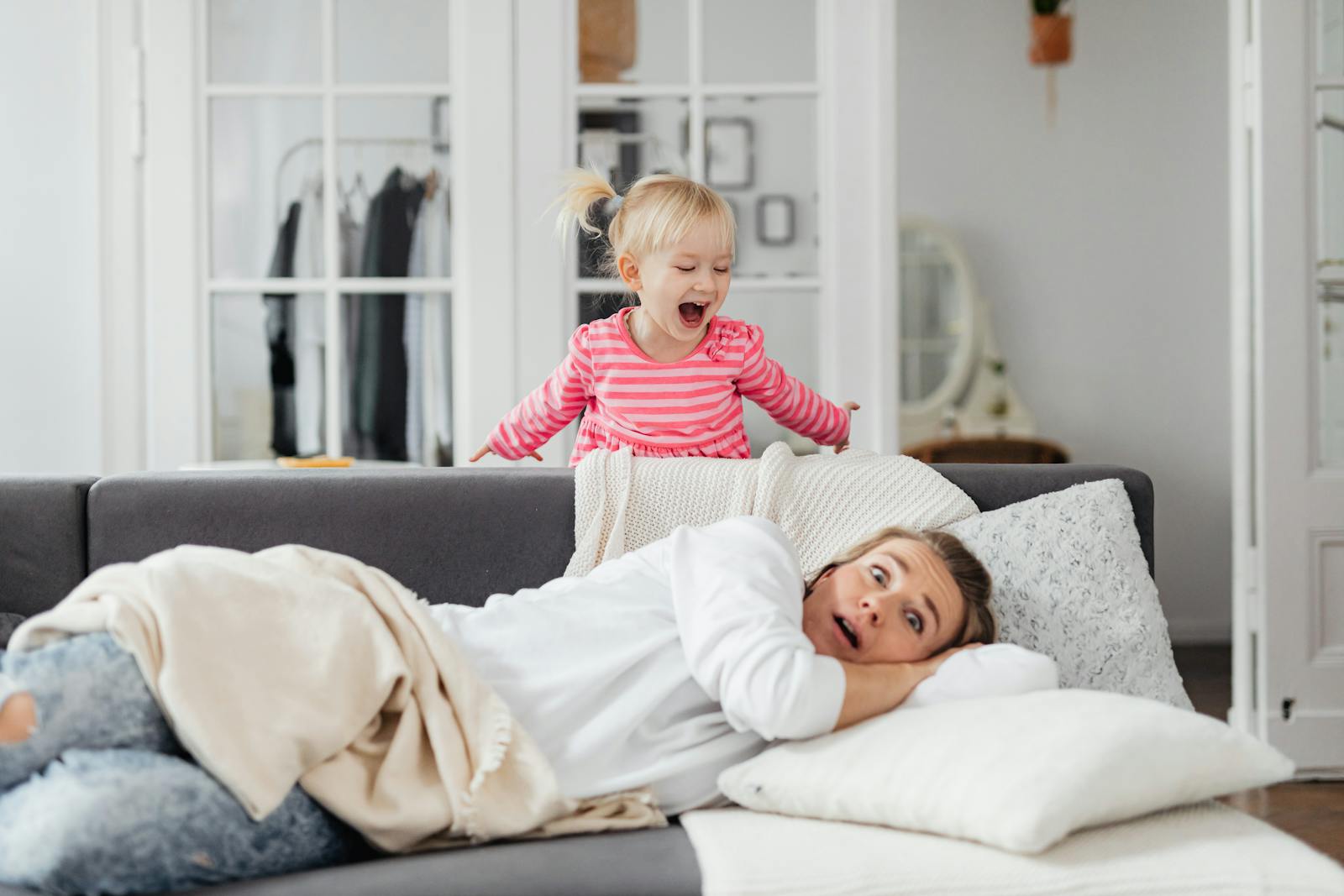 A young girl playfully shouting behind a resting mother on a couch in a cozy room.