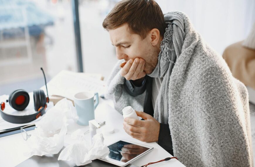 Man wrapped in blanket feeling unwell at home, surrounded by tissues and medication.