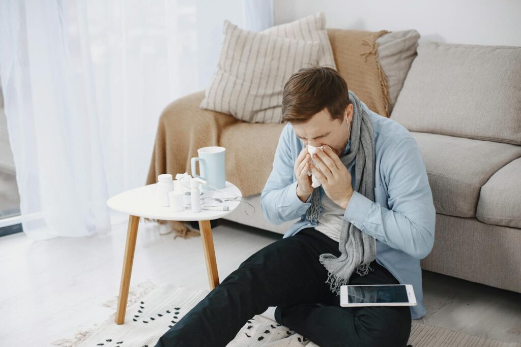 A man in a living room sneezing into a tissue, surrounded by a table, sofa, and digital tablet.