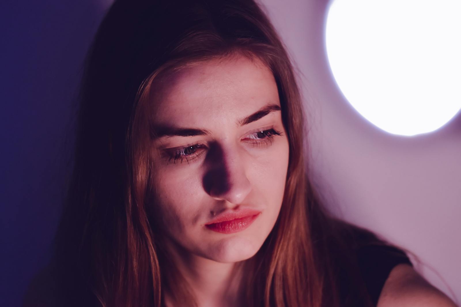 A young woman with long hair gazes thoughtfully, illuminated by gentle lighting indoors.