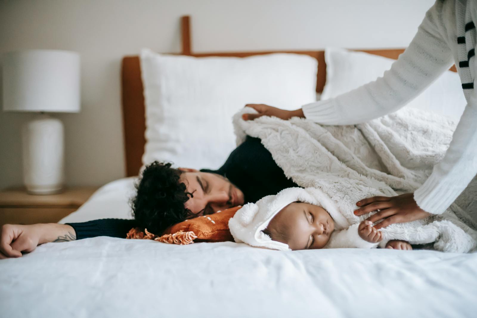 Crop faceless black woman in casual clothes spreading blanket on diverse husband and baby sleeping together on soft bed in morning