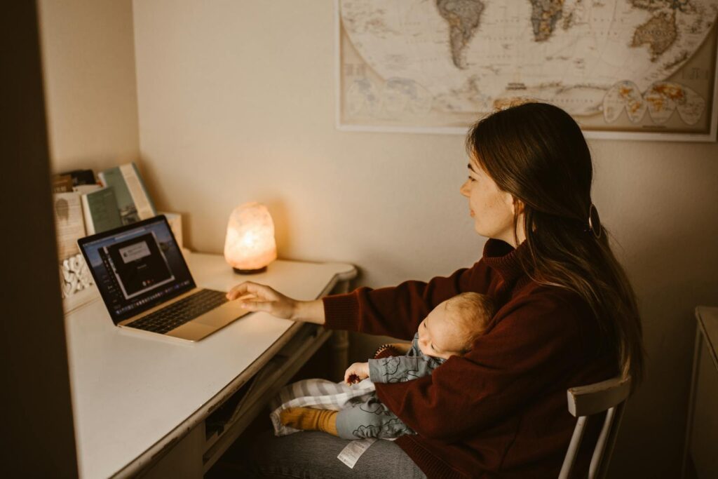A mother working on a laptop while holding her sleeping baby, creating a warm at-home atmosphere.
