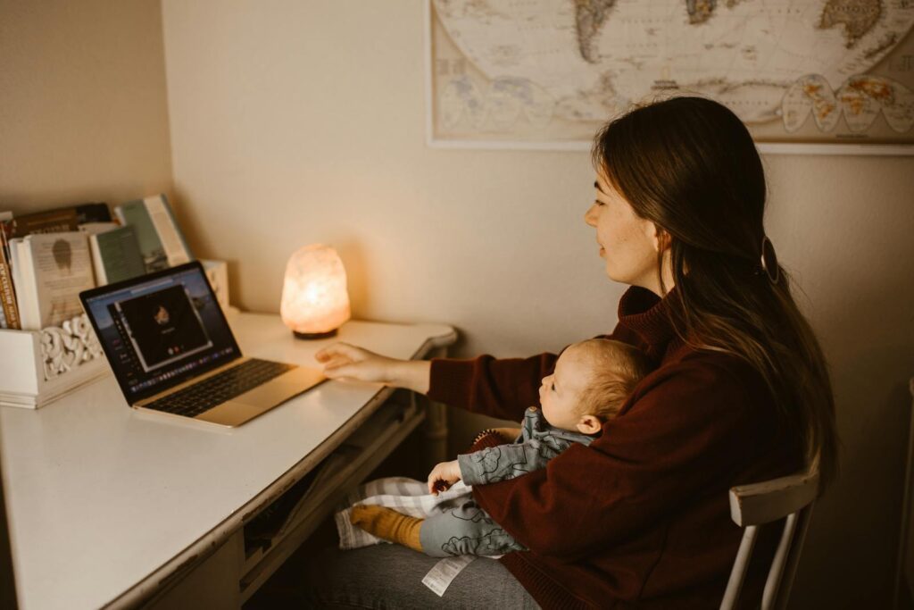 A mother multitasking with a baby on her lap while video calling from a desk at home.
