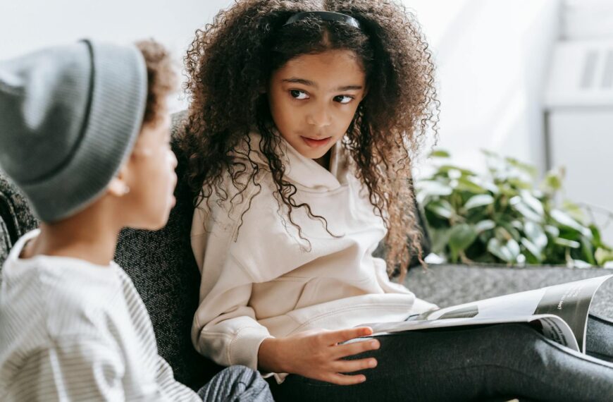Crop African American smart cute girl with curly hair reading interesting magazine for anonymous brother in daytime