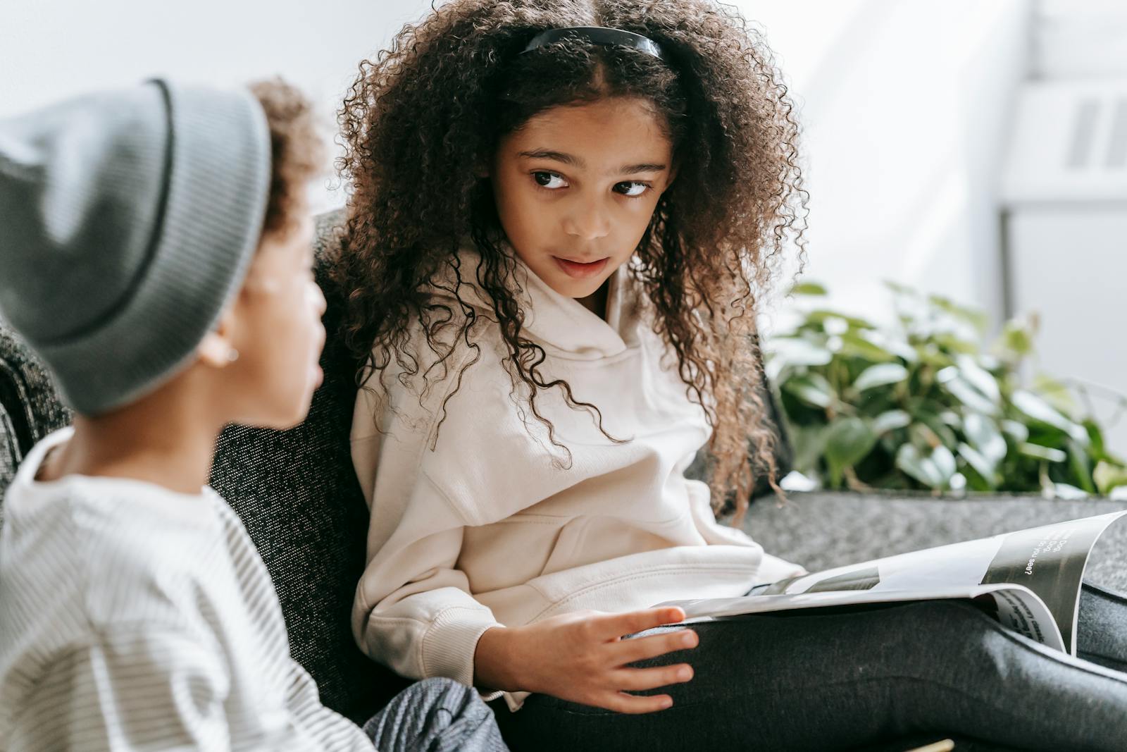 Crop African American smart cute girl with curly hair reading interesting magazine for anonymous brother in daytime