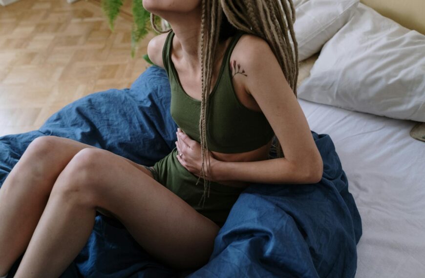 Woman sitting in bed holding her stomach, indicating pain or discomfort, possibly due to cramps.