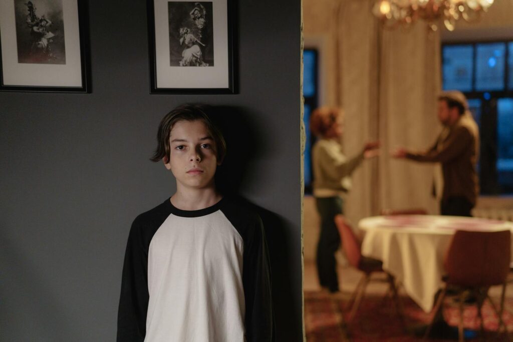 A boy stands in focus, while two adults argue in the background, depicting family conflict.