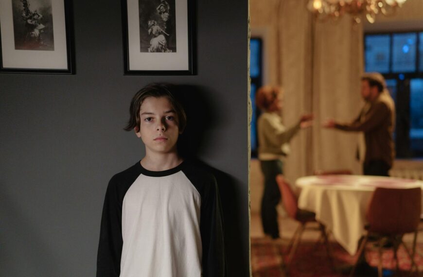 A boy stands in focus, while two adults argue in the background, depicting family conflict.