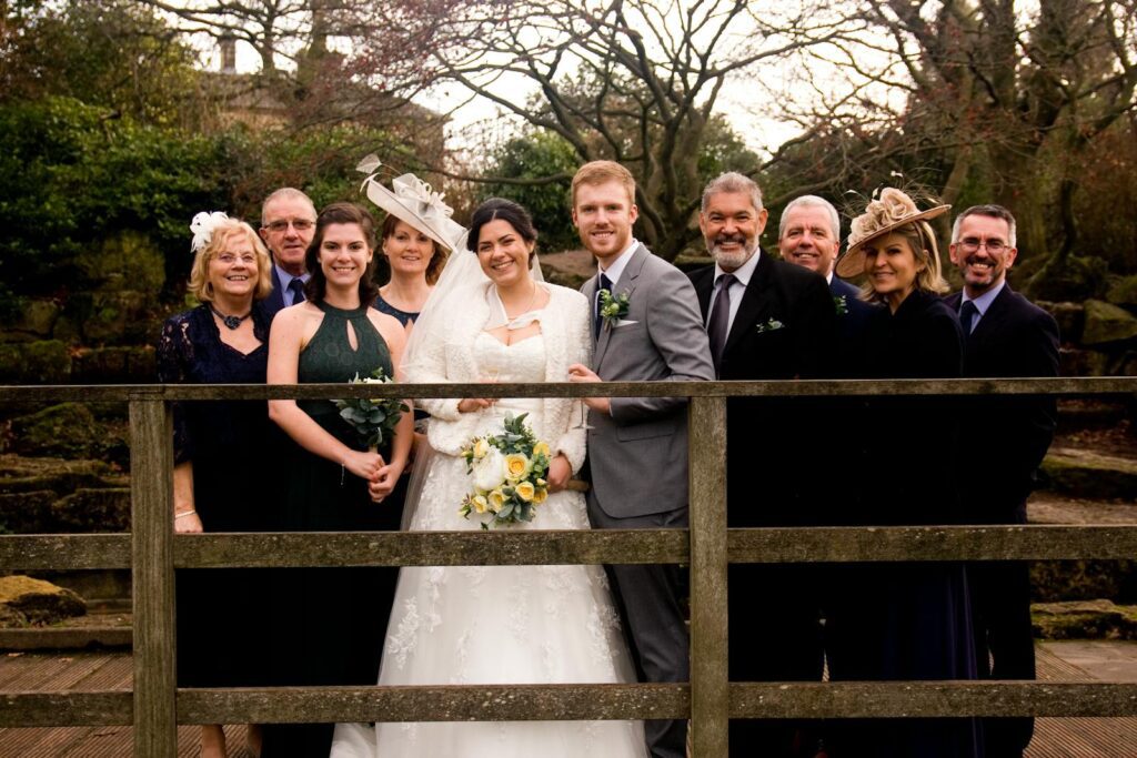Outdoor wedding group photo featuring bride, groom, and family on a rustic wooden bridge.