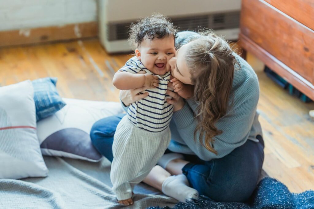 Full length caring mother in casual wear hugging and kissing cute ethnic baby cheek while sitting on floor in light living room