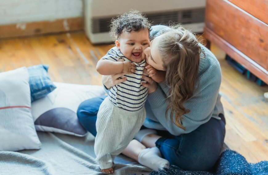 Full length caring mother in casual wear hugging and kissing cute ethnic baby cheek while sitting on floor in light living room
