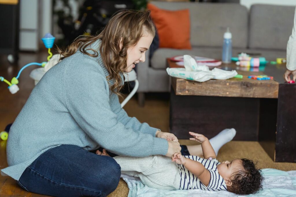 Full body cheerful young mother playing with and putting pants on cute ethnic baby lying on floor at home