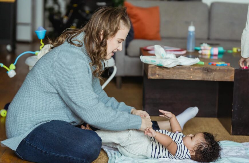 Full body cheerful young mother playing with and putting pants on cute ethnic baby lying on floor at home