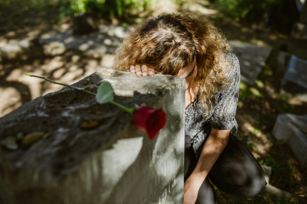 A woman grieves at a cemetery, resting her head on a tombstone with a rose, capturing deep emotion.