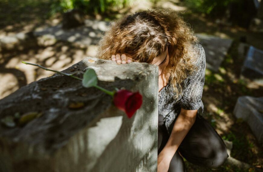 A woman grieves at a cemetery, resting her head on a tombstone with a rose, capturing deep emotion.