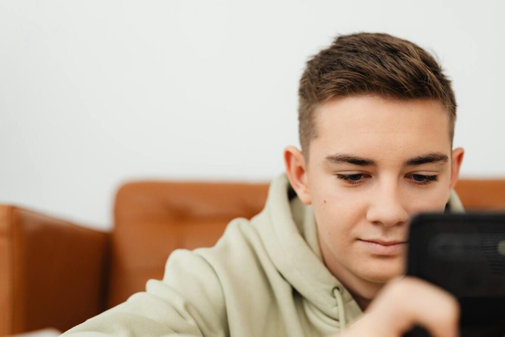 Close-up of a teenager using his mobile phone, sitting on a brown sofa in casual attire.