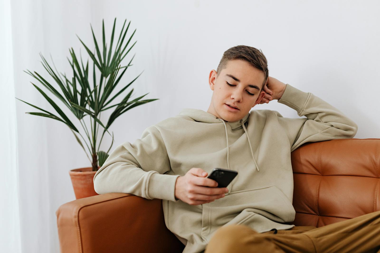 Teenage boy sitting indoors texting on a smartphone, wearing a hoodie.