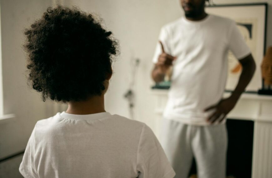 Back view of unrecognizable black boy with Afro hair standing in front of crop anonymous dad pointing finger and scolding at home