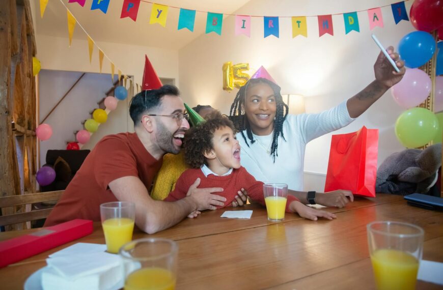 Happy family celebrating a child's birthday with balloons and party hats, capturing a joyful moment with a selfie.