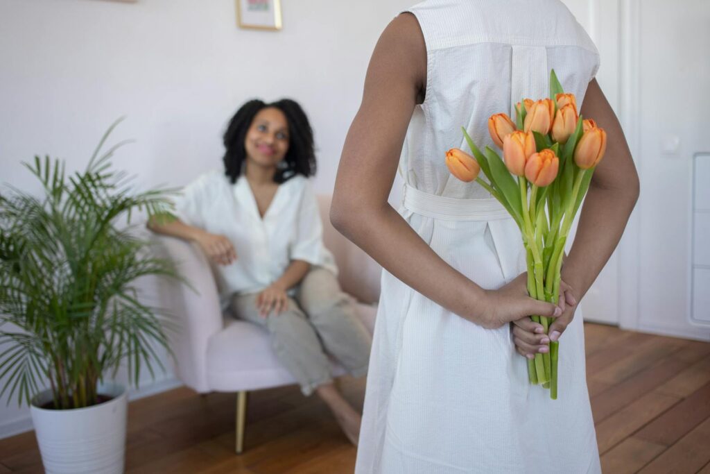 A young girl surprises her mother with a bouquet of orange tulips indoors. Cozy and heartwarming moment.