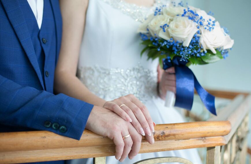 Bride and groom in elegant attire with bouquet and rings on wooden railing.