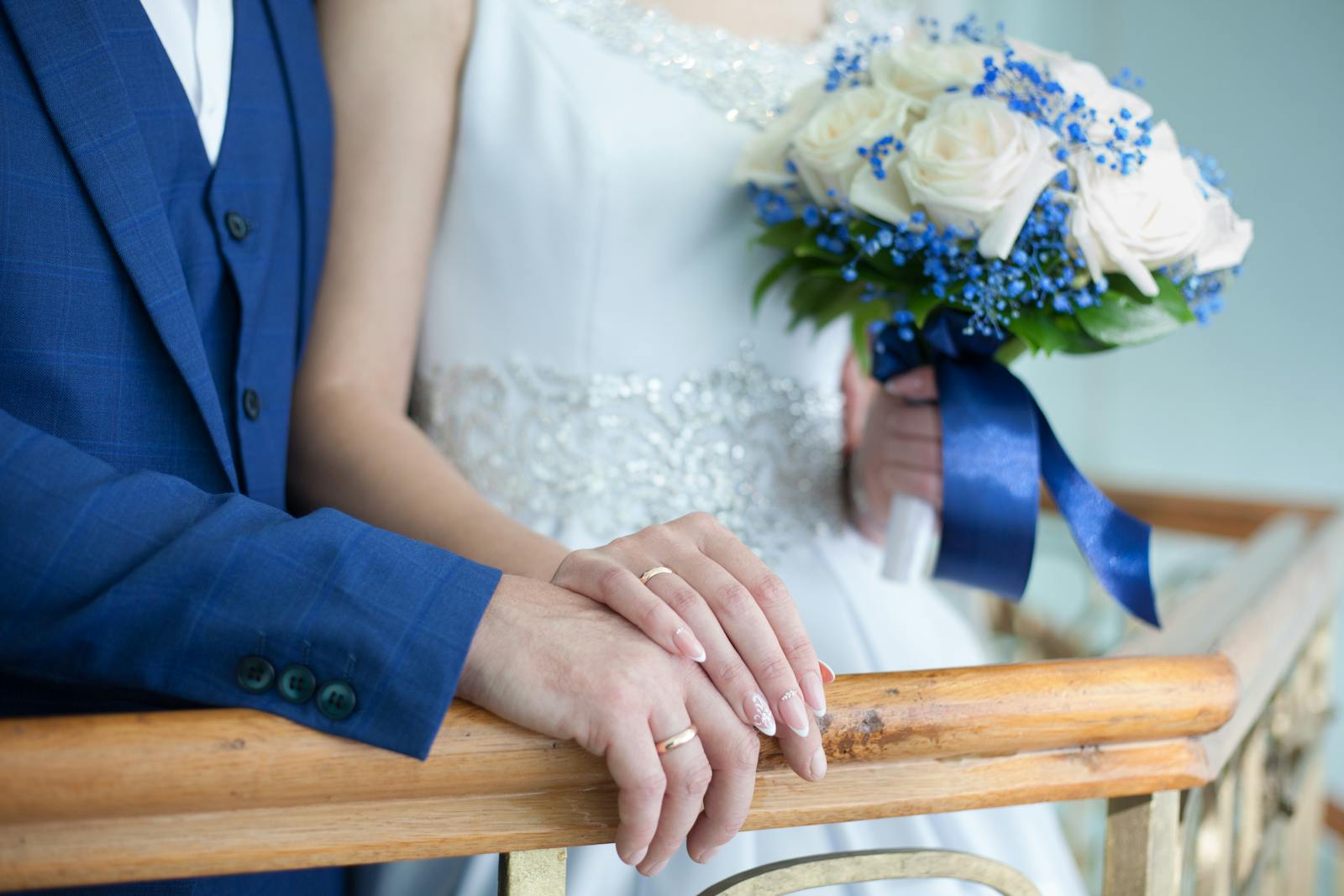 Bride and groom in elegant attire with bouquet and rings on wooden railing.